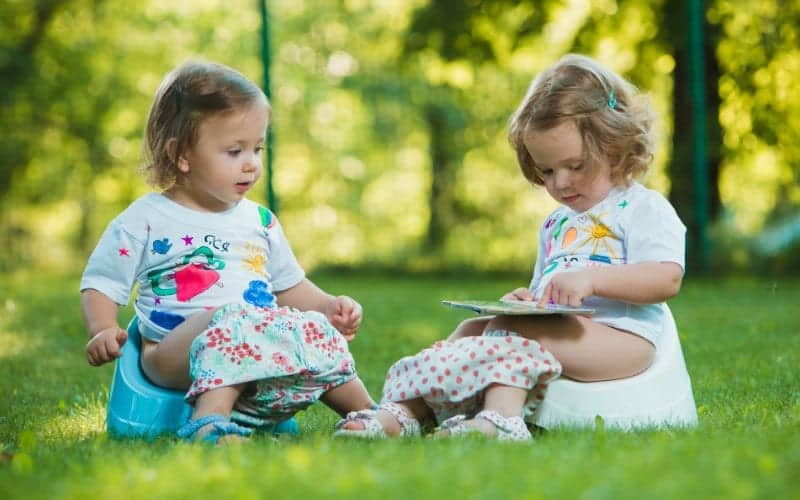 two toddlers getting potty trained sitting on the grass with potty training supplies